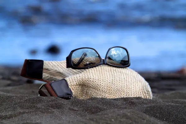 Eine Sonnenbrille liegt auf einem Strohhut, der im Sand am Strand platziert ist, während das Wasser im Hintergrund glitzert.