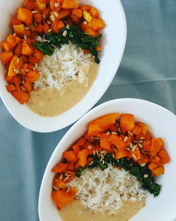 Veganes Kürbis Süßkartoffel Curry liegt in zwei Schalen, umgeben von Reis und Erdnusssauce, appetitlich angerichtet.
