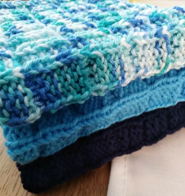 Bunte, gestrickte Spültücher liegen übereinander auf einem Holzuntergrund und zeigen verschiedene Blautöne.