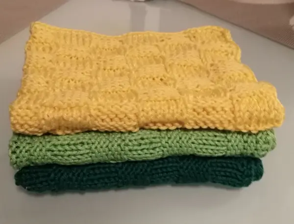 Gestapelte, handgestrickte Spültücher in Gelb, Grün und Dunkelgrün liegen auf einer glatten Oberfläche.