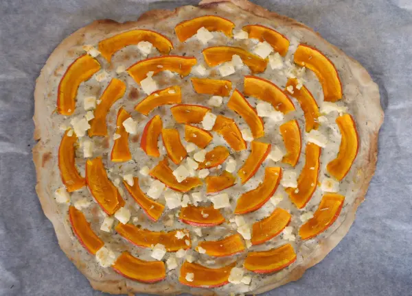 Ein runder Flammkuchen mit Kürbis und Feta ist frisch auf einem Backblech platziert und bereit zum Backen.