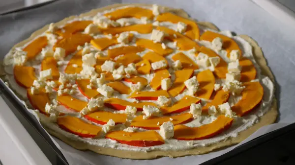 Ein Flammkuchen mit Kürbis und Feta liegt auf einem Blech, bereit zum Backen in einer modernen Küche.
