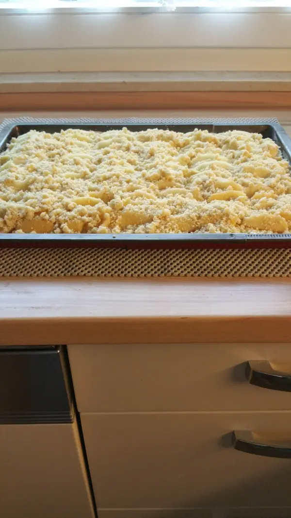 Ein Blech voller saftigem Apfelkuchen mit Streuseln steht auf einer Arbeitsplatte in einer modern eingerichteten Küche.
