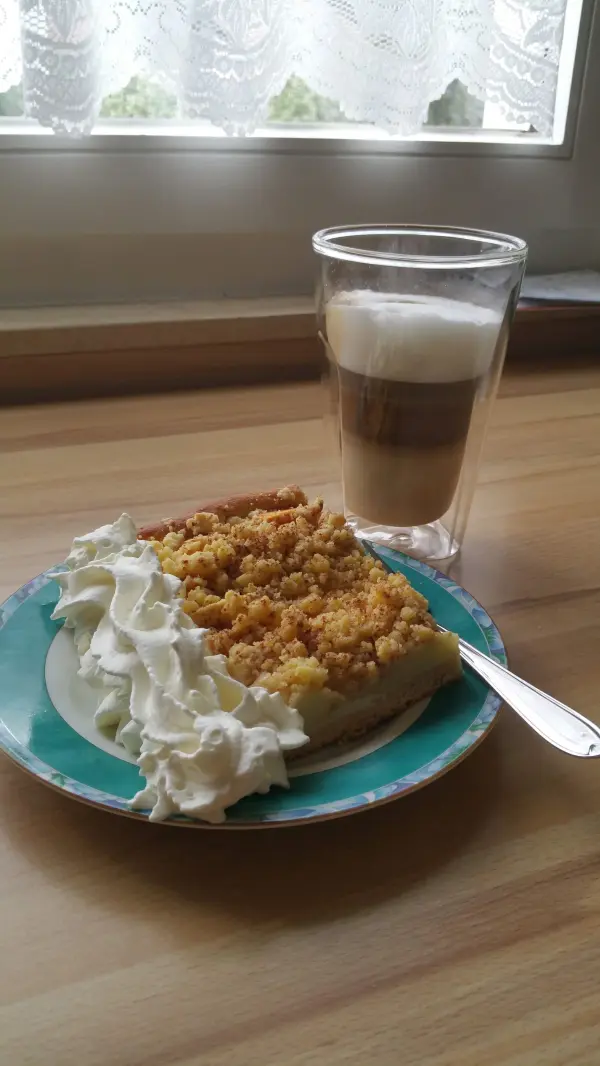 Ein saftiges Stück Apfelkuchen mit Streuseln wird auf einem Teller mit Sahne serviert, daneben steht ein Glas Kaffee.
