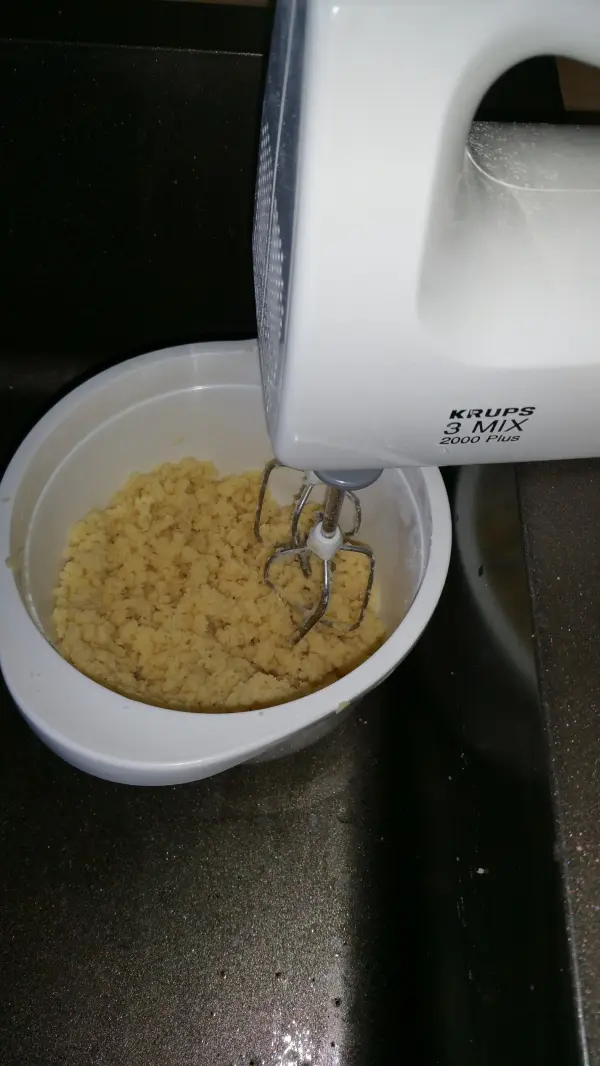 Ein Handmixer rührt in einer weißen Schüssel Grießbrei, der für einen saftigen Apfelkuchen zubereitet wird.