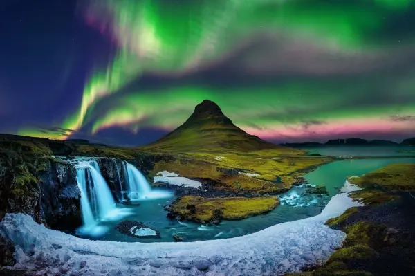 Ein Wasserfall fließt in einen glitzernden Fluss, während das Nordlicht über einem schneebedeckten Hügel leuchtet.