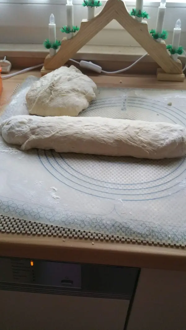 Teigwege werden vorbereitet; ein geformter Brötchenteig liegt auf einer Arbeitsfläche in einer hellen Küche.