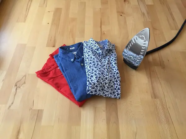 Ein Dampfbügeleisen steht neben einem Stapel gefalteter Kleidung in Rot, Blau und mit Muster auf einem Holzboden.