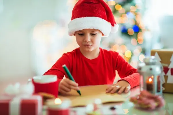 Ein Kind trägt einen Weihnachtsmann-Hut und schreibt konzentriert an einem Tisch mit Geschenken und Kerzen.