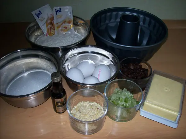 Eine Guglhupf-Form steht im Mittelpunkt, umgeben von Zutaten wie Eiern, Mehl und Butter zur Zubereitung von Rührteig.