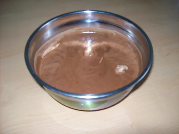 Eine silberne Schüssel enthält eine cremige Mousse au Chocolat, verdeckt von einem leicht glänzenden Film, auf einem hellen Tisch.