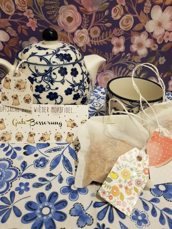 Ein dekorativer Teekessel steht neben einer Tasse auf einem blühenden Tischdeckenmuster, umgeben von hübschen Teebeuteln.