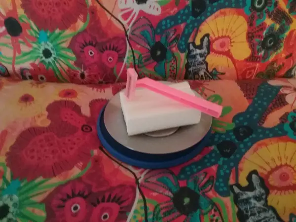 Ein pinker Rasierer liegt auf einem weißen Seifenstück, platziert auf einem runden blauen Untersetzer, vor einem bunten, floralen Hintergrund.