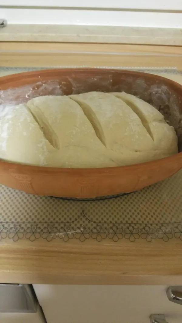 Ein runder Joghurtbrotteig liegt in einem Römertopf, bereit zum Backen, auf einer Holzoberfläche in heller Umgebung.