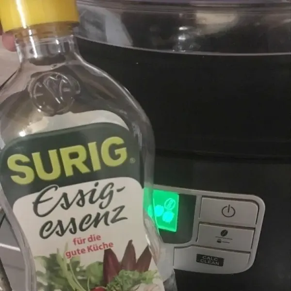 Eine Flasche Essig-Essenz steht neben einer Kaffeemaschine, die grün leuchtet, zum Entkalken und Reinigen.