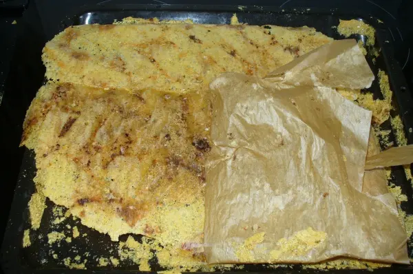 Käsechips liegen auf einem Backblech, leicht verbrannt und teilweise zerbrochen, umgeben von Käsespänen und Pergamentpapier.