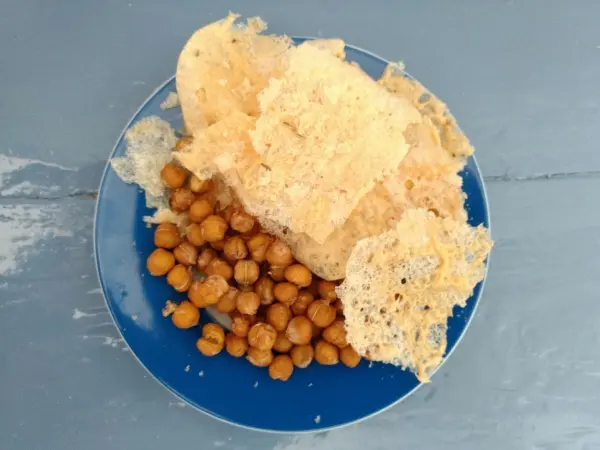 Käsechips liegen knusprig auf einem blauen Teller, umgeben von kleinen, gerösteten Kichererbsen auf einem grauen Hintergrund.