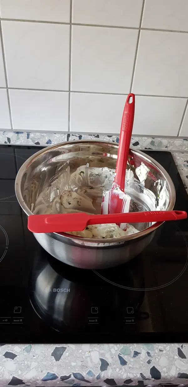 Ein roter Teigschaber aus Silikon steht in einer Schüssel mit Mischung, platziert auf einem Herd mit Fliesen.