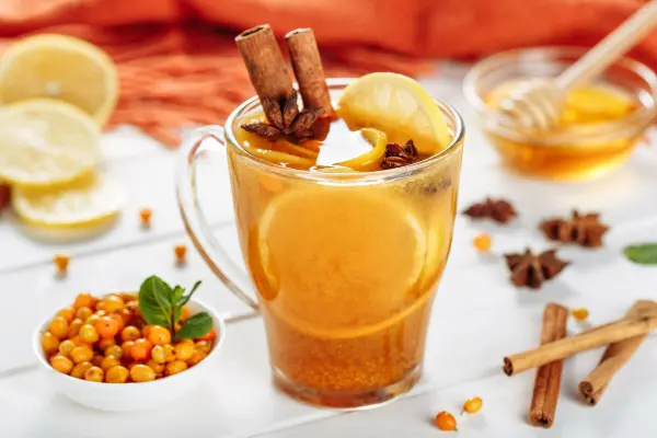 Ein Glas Sanddorntee mit Ingwer und Zimt steht auf dem Tisch, umgeben von Zitrone, Honig und Beeren, in einem hellen Ambiente.