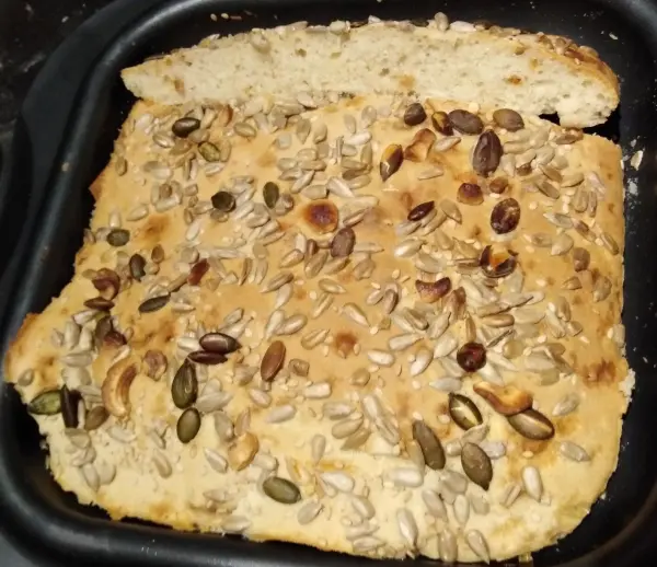 Ein Fladenbrot mit einer goldbraunen Oberfläche, belegt mit Sonnenblumenkernen und Kürbiskernen, liegt in einer schwarzen Form.