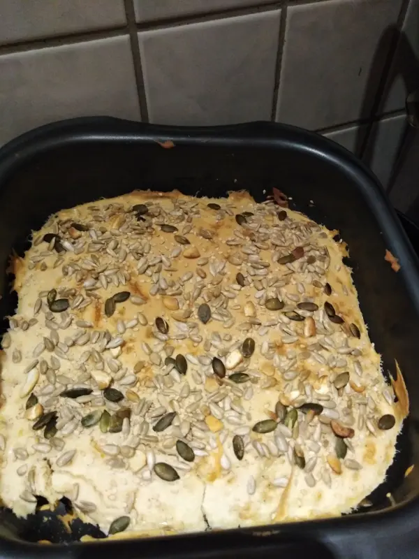 Ein Fladenbrot mit einer goldbraunen Oberfläche, belegt mit verschiedenen Samen, liegt in einer schwarzen Backform auf einer Küchenoberfläche.