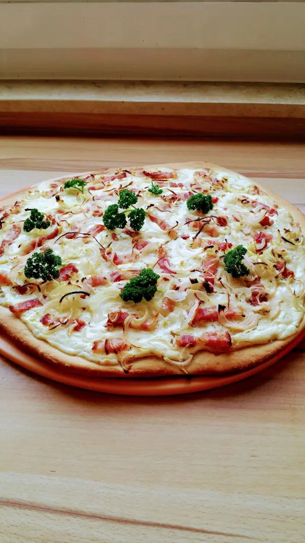 Ein frisch gebackener Flammkuchen liegt auf einem Holzbrett. Er ist belegt mit Speck, Zwiebeln und frischen Kräutern.