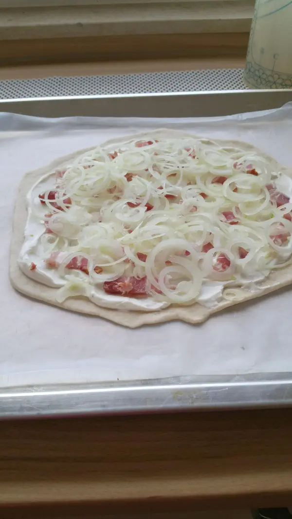 Ein belegter Elsässer Flammkuchen liegt auf einem Backblech, garniert mit Zwiebeln und einer cremigen Schicht.