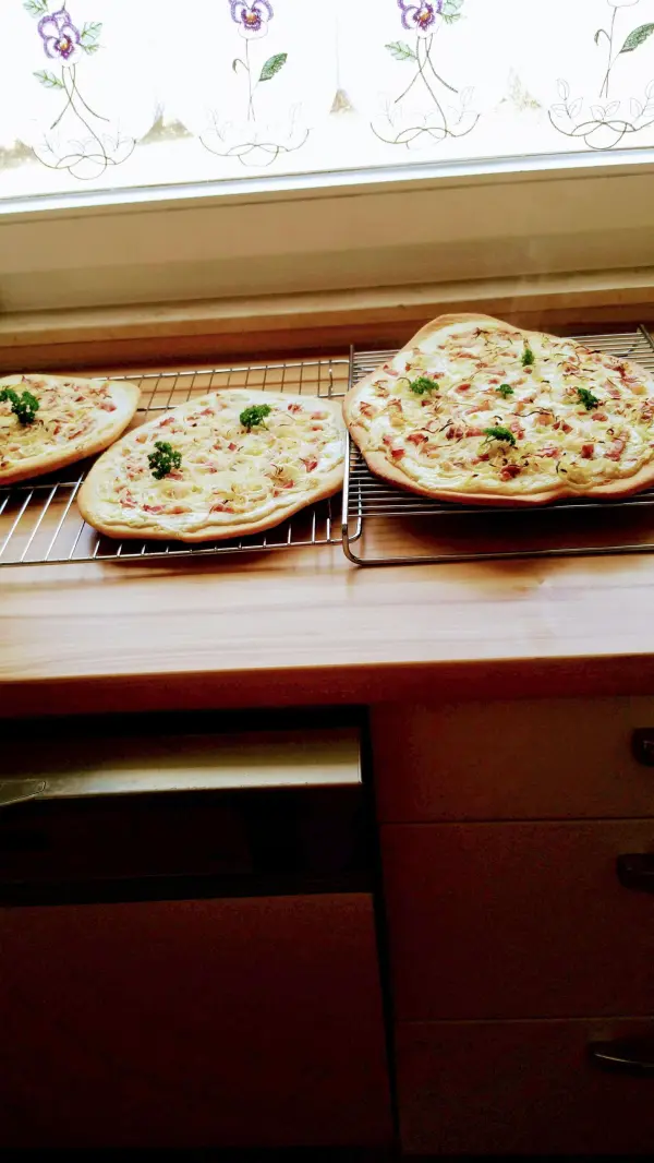Drei frisch gebackene Elsässer Flammkuchen liegen auf einem Kühlregal in einer hellen Küche.