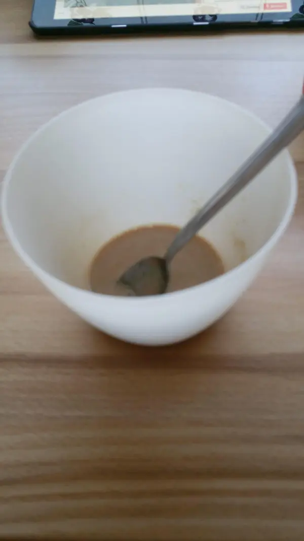 Eine leere weiße Schüssel steht auf einem Holztisch, in der ein wenig braune Flüssigkeit mit einem Löffel vermischt wird.