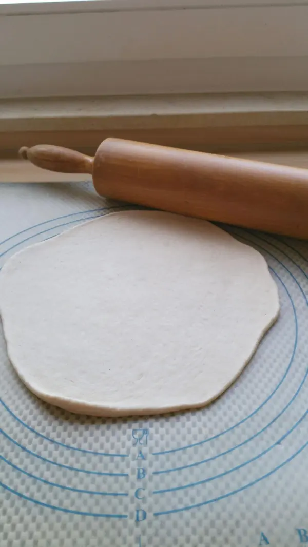 Ein frisch ausgerollter Flammkuchenteig liegt auf einer. Arbeitsfläche, daneben liegt ein Holzrollingpin.