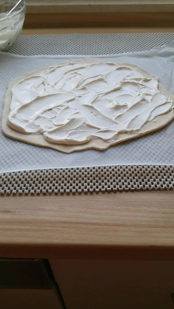 Ein runder Flammkuchenteig liegt auf einer Arbeitsfläche, gleichmäßig mit einer cremigen Schicht bedeckt, bereit zum Belegen.