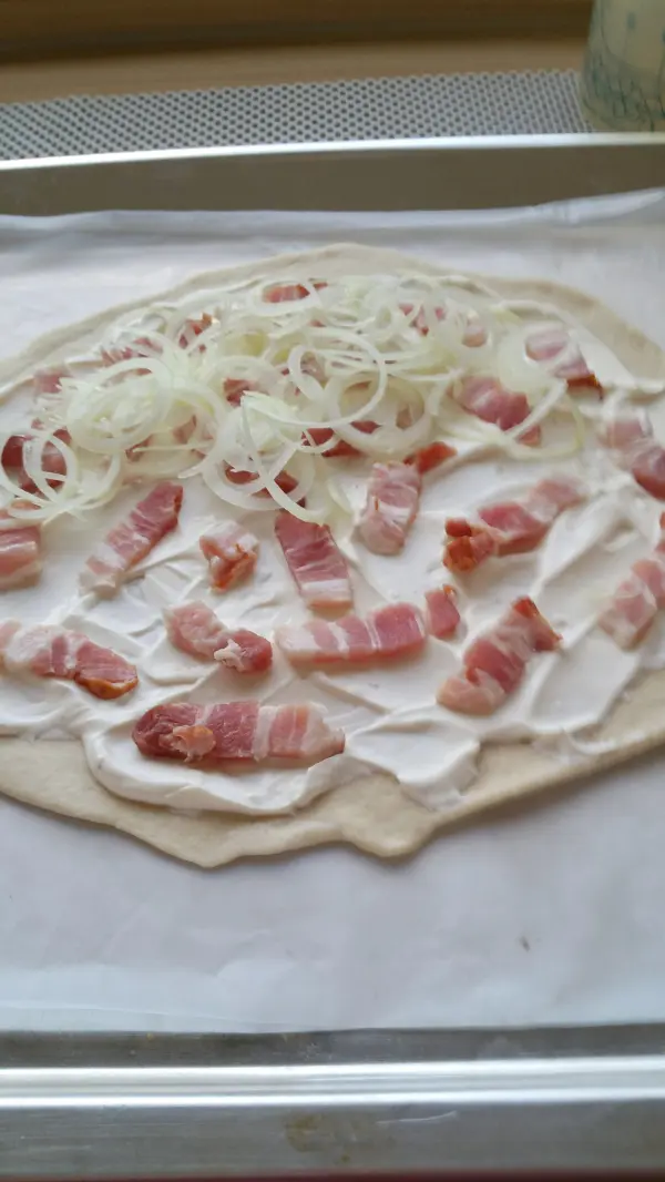 Ein Flammkuchen wird auf einem Backblech vorbereitet, belegt mit Speck und Zwiebeln auf einem dünnen Teig.
