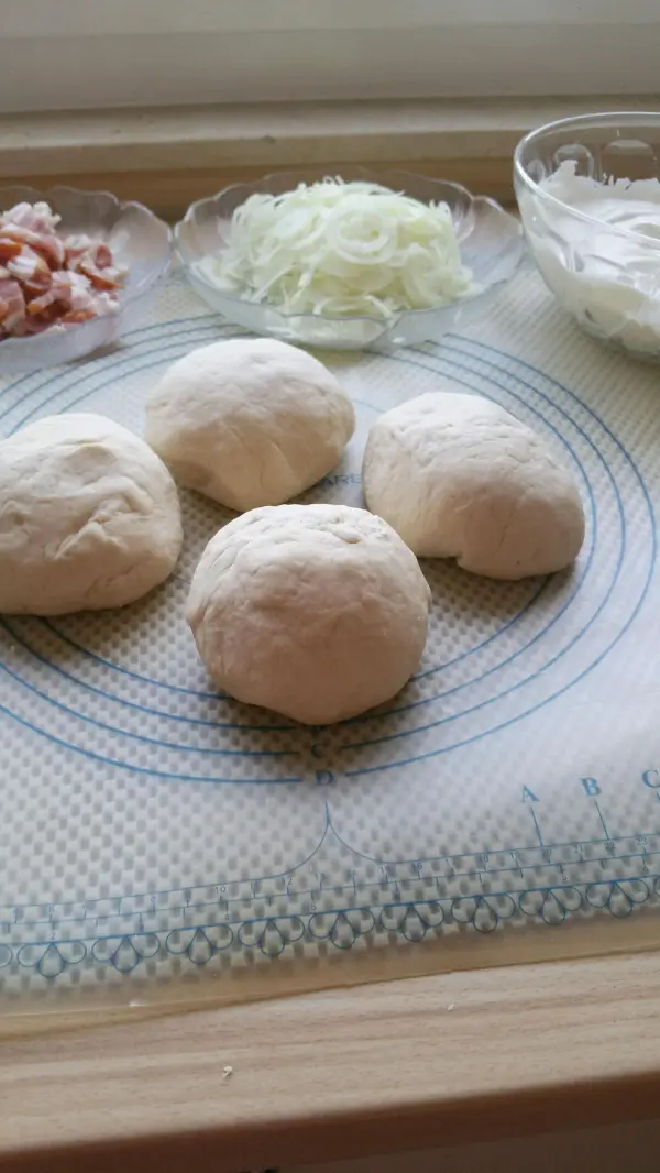 Vier Teigbällchen liegen auf einer Arbeitsfläche, umgeben von Zutaten wie Zwiebeln, Speck und Creme, bereit für Elsässer Flammkuchen.