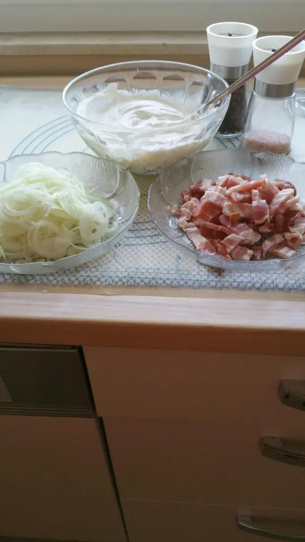 Ein frisch zubereiteter Elsässer Flammkuchen hat Schalen mit Zwiebeln, Speck und einer cremigen Sauce auf einem Tisch platziert.