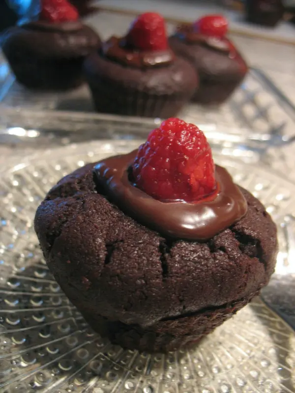Schoko-Himbeer-Muffins stehen auf einem Glasteller, glasiert mit Schokolade und dekoriert mit einer Himbeere obenauf.