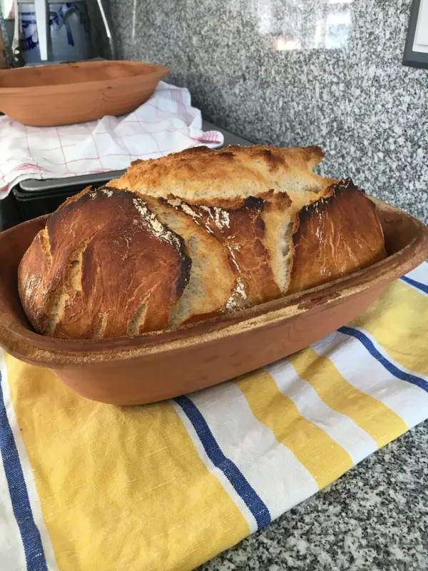 Ein frisch gebackenes Dinkel-Roggenbrot liegt in einem Römertopf, platziert auf einem gestreiften Geschirrtuch in einer modernen Küche.