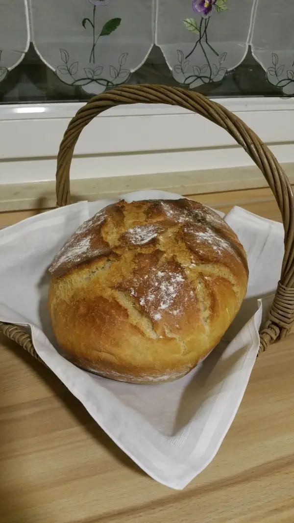 Ein frisch gebackenes Dinkel-Roggenbrot liegt in einem Korb, umgeben von einem weißen Tuch auf einer Holzoberfläche.