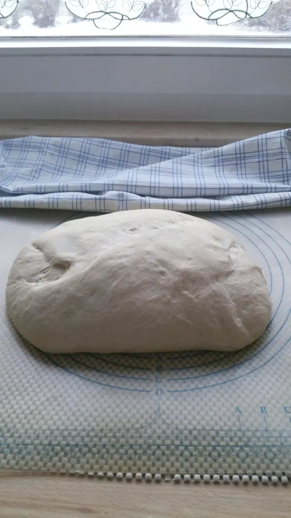 Ein gehobener Brotteig liegt auf einer Backunterlage, während ein Küchentuch darüber drapiert ist, bereit zum Dinkel-Roggenbrot-Backen.
