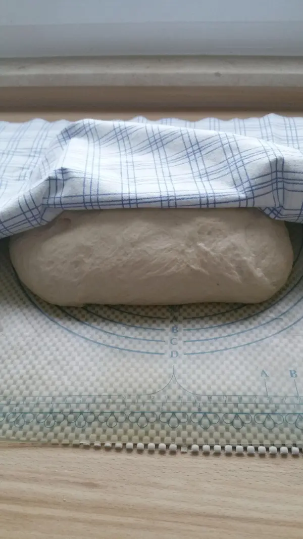 Der Brotteig liegt unter einem karierten Küchentuch auf einer Backunterlage und ruht vor dem Backen im Römertopf.