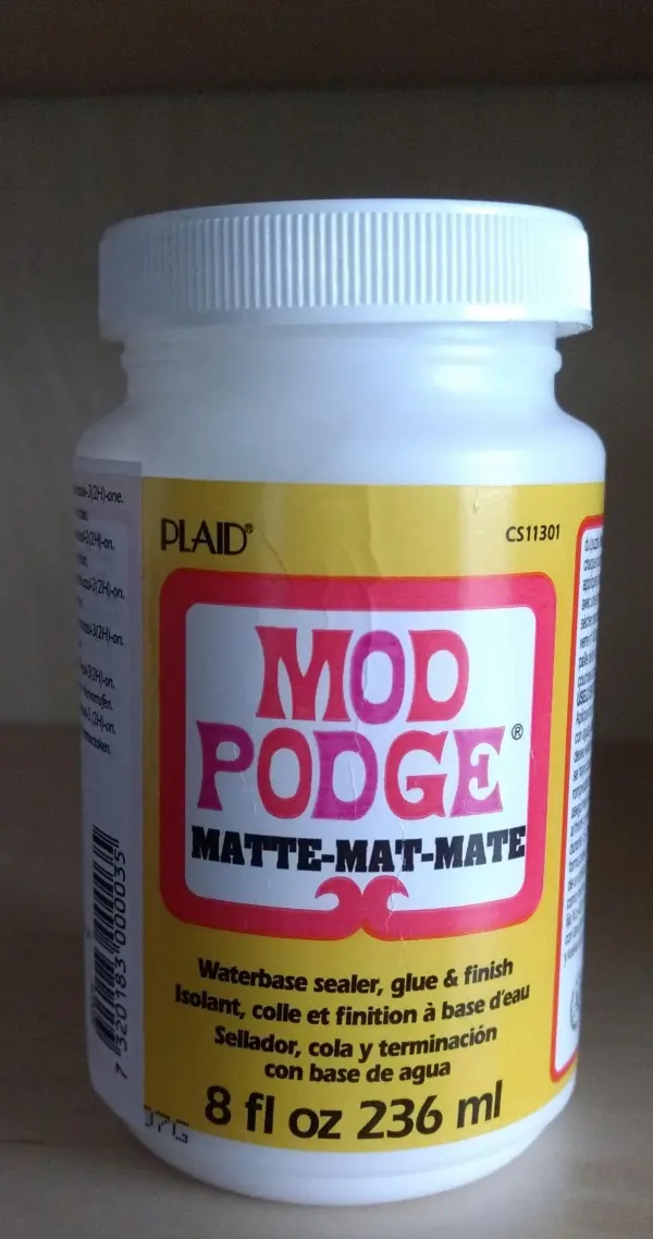 Ein Behälter mit Mod Podge steht auf einem hellen Regal. Er wird verwendet, um ausgeblasene Eier im Zeitungslook zu verzieren.