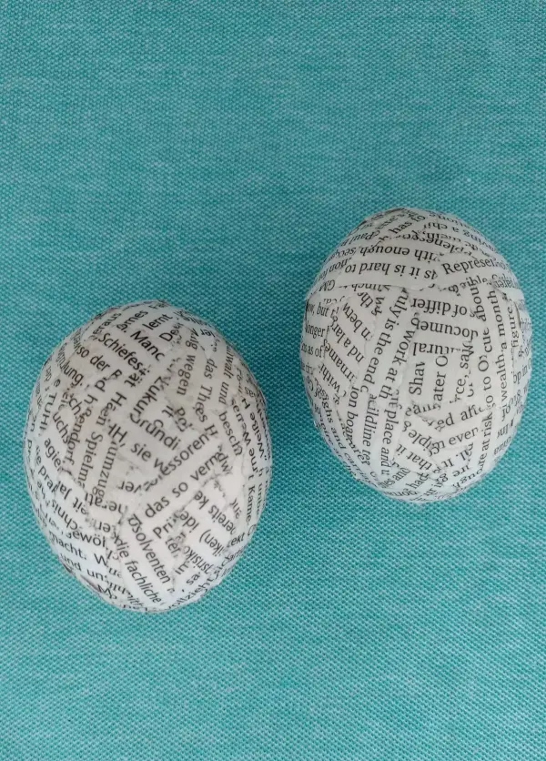 Ausgeblasene Eier sind mit Zeitungsdruck bedruckt und liegen auf einem sanften, türkisfarbenen Hintergrund.