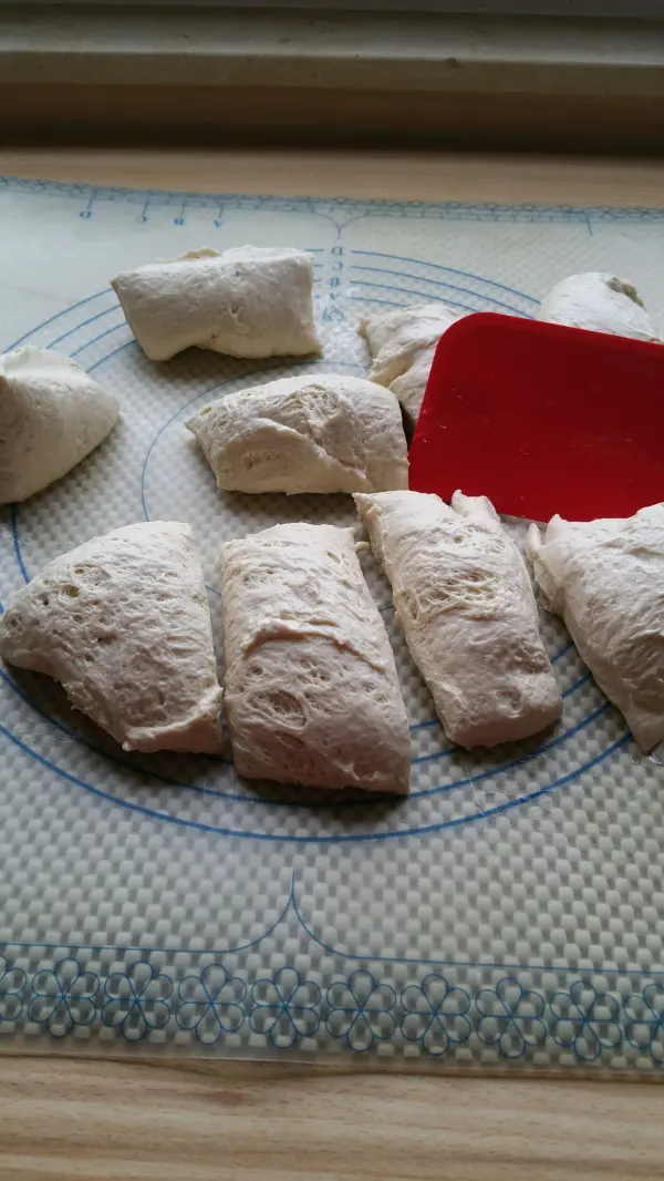Auf einer bemehlten Unterlage liegen frisch geformte Joghurtbrötchen, bereit zum Backen, neben einem roten Spatel.