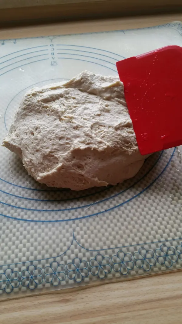 Ein Joghurtteig liegt auf einer Unterlage, während ein roter Spatel ihn bearbeitet, um Brötchen vorzubereiten.