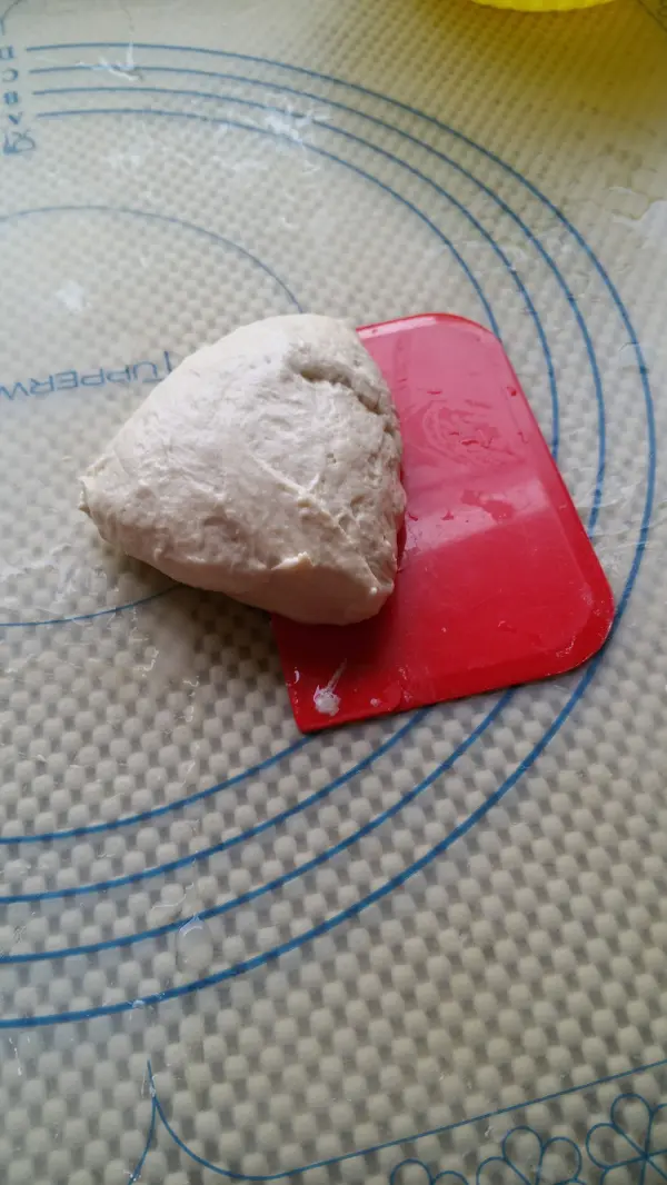 Ein frischer, weißer Teig für Joghurtbrötchen liegt auf einer Arbeitsfläche, während ein roter Teigschaber daneben liegt.