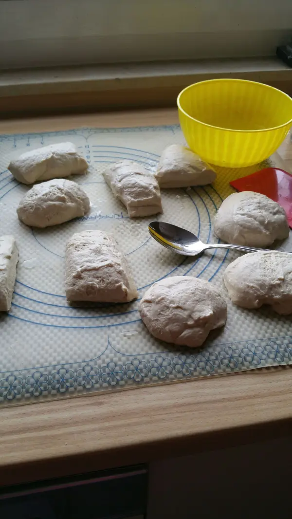 Auf einem Backbrett liegen mehrere ungebackene Joghurtbrötchen, die bereit sind, gebacken zu werden, neben einer gelben Schüssel.