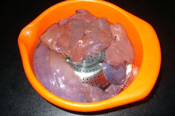Frische Leber liegt in einem Sieb in einer orangefarbenen Schüssel und lässt abtropfen, auf einer dunklen Oberfläche.