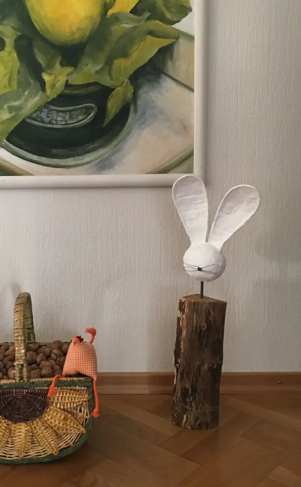 Ein weißer Osterhase aus Papier sitzt auf einem Holzstück in einem gemütlichen Wohnraum neben einem geflochtenen Korb.