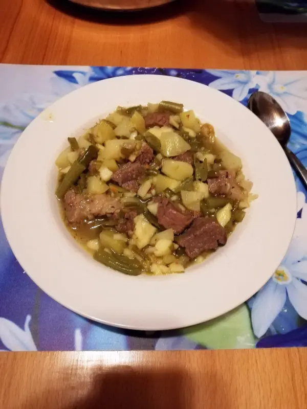 Ein Bohneneintopf mit Äpfeln befindet sich in einer weißen Schüssel, gefüllt mit Gemüse und Fleischstücken, auf einem hellen Tisch.