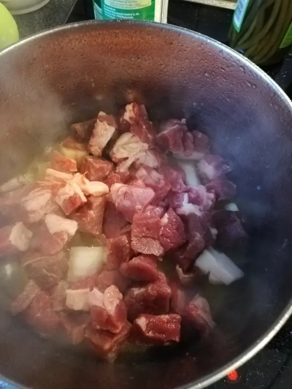 Im Topf braten Stücke von Fleisch und Zwiebeln, während Dampf aufsteigt und eine warme Küche umgibt.