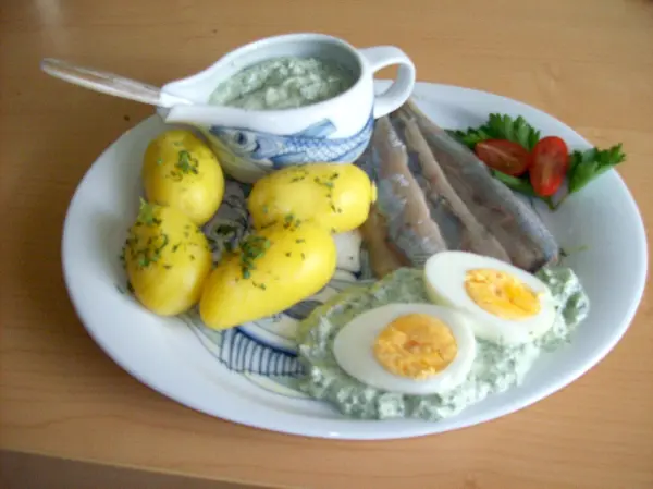 Auf dem Teller liegen gekochte Kartoffeln, frische Matjes und Eier, begleitet von einer Schüssel grüner Soße.